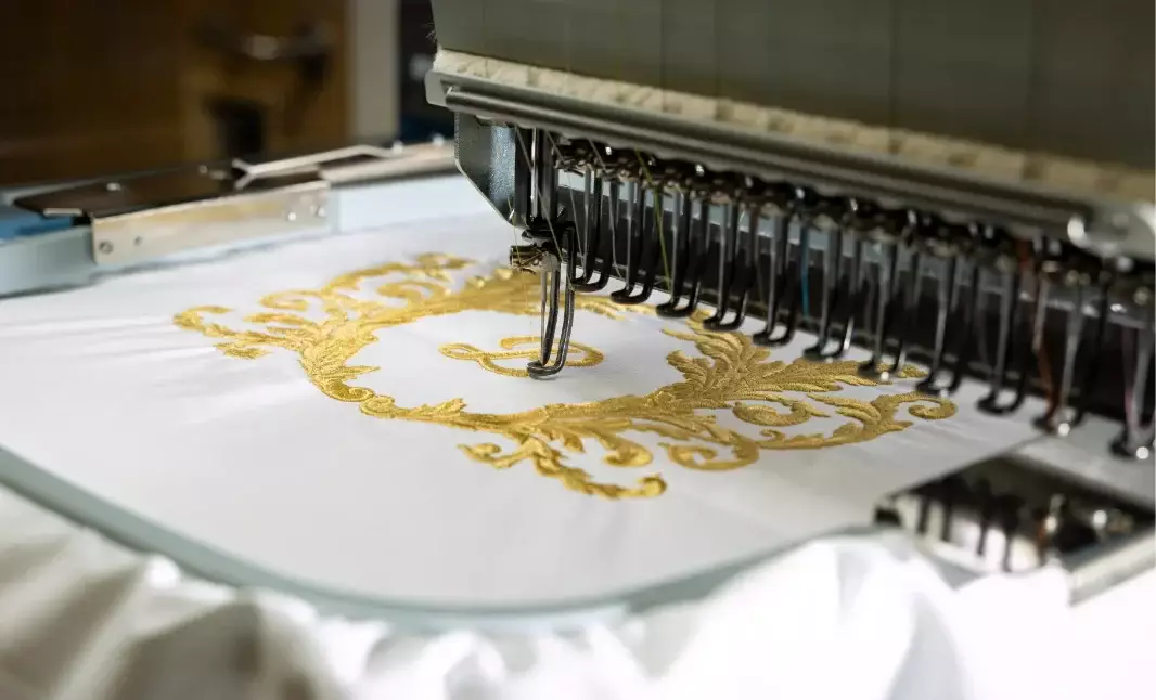 Embroidery machine stitching an ornate gold design onto white fabric, highlighting bespoke detailing and premium textile craftsmanship.